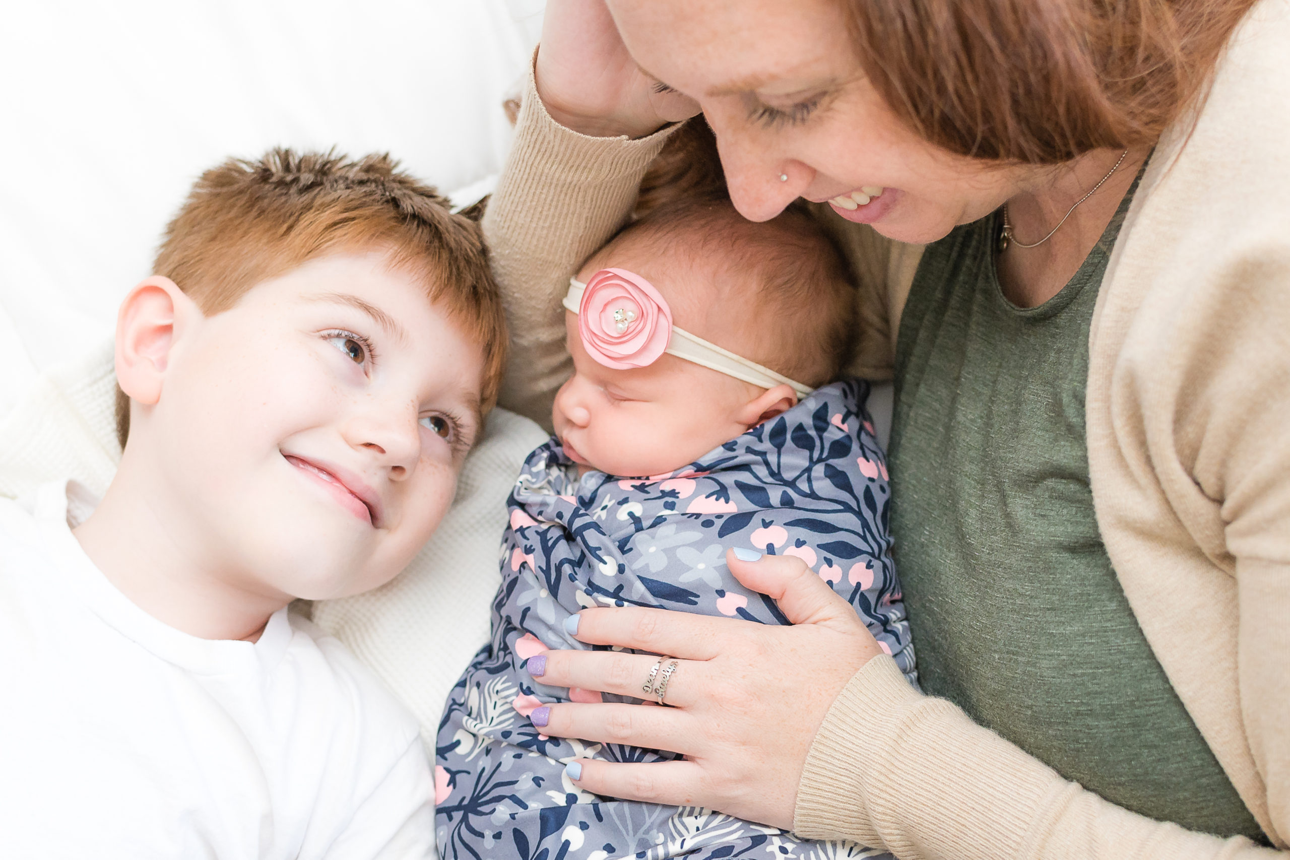 mother, son, and newborn daughter are laying on the bed during mommy and me lifestyle newborn session. son is looking up at mother smiling while mother is looking down at her two children.